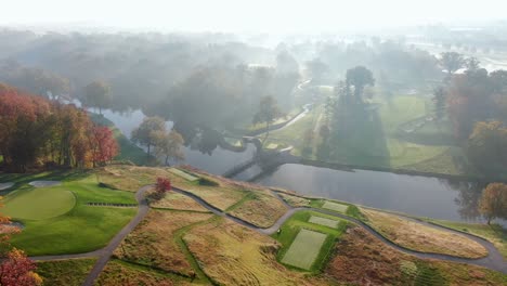 Vista-Aérea-Del-Amanecer-Sobre-El-Campo-De-Golf-En-La-Mañana-Brumosa,-La-Luz-Espectacular-Cae-Sobre-Las-Hojas-De-Los-árboles-De-Otoño,-El-Río,-El-Puente