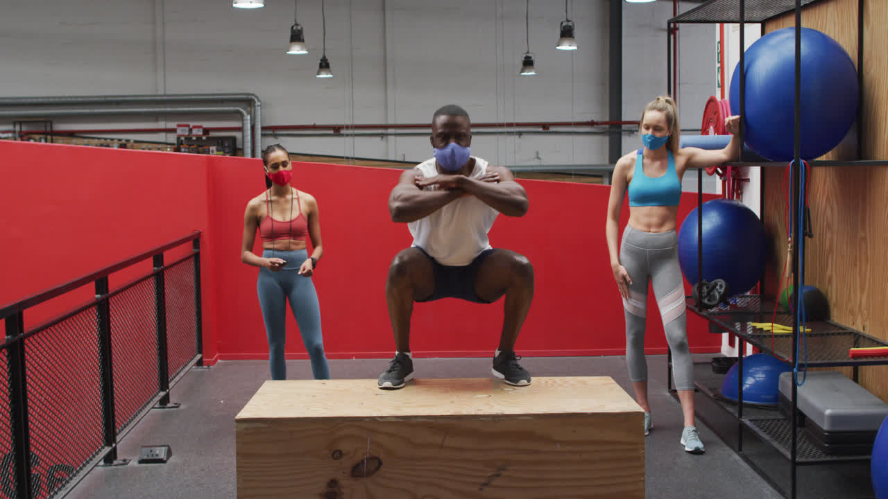 아프리카계 미국인 남자가 체육관에서 얼굴 마스크를 쓰고 상자에 뛰어다니고, 두 명의 다양한 여성이 지켜보고 있습니다.