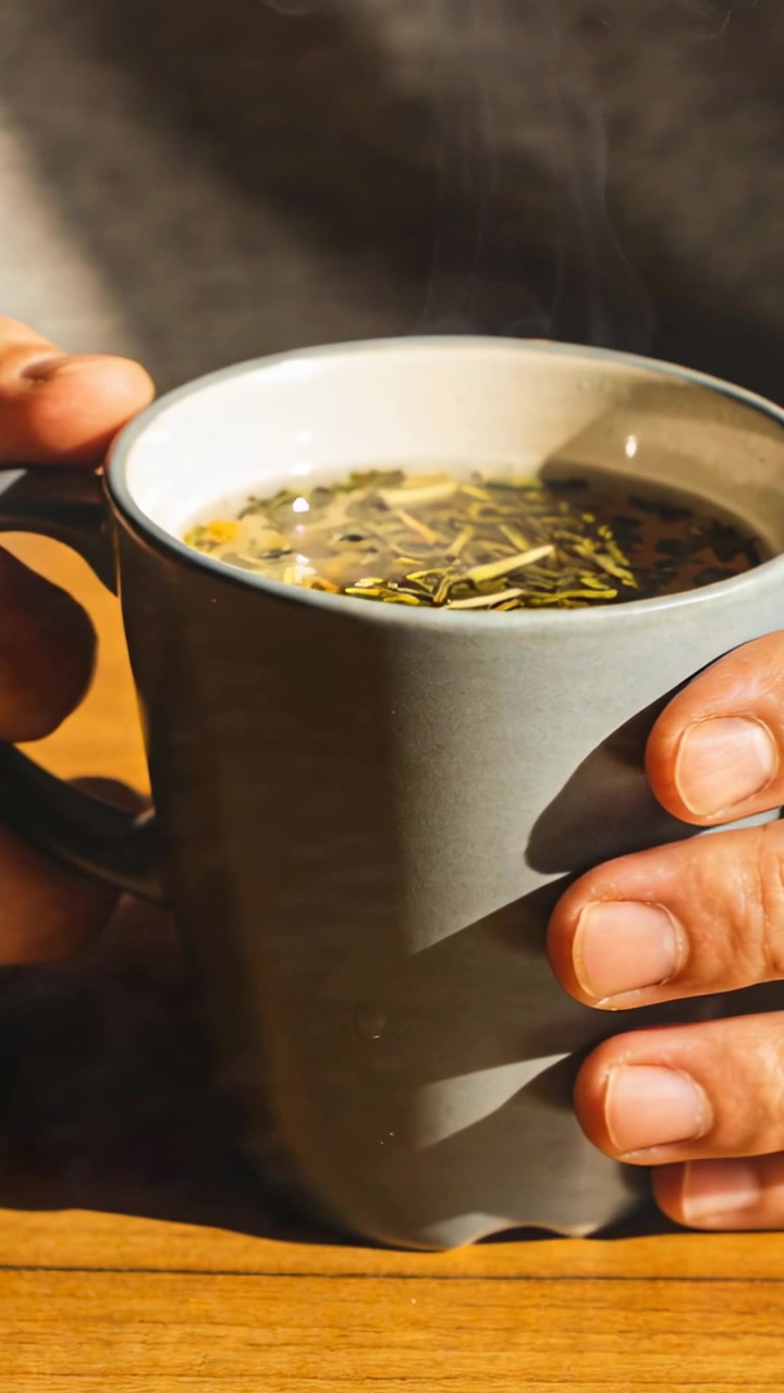 Video verticale: Si nota del vapore, mani con polsino grigio che tengono ferma una tazza su un tavolo di legno con erbe aromatiche sparse.
