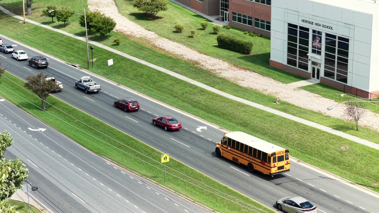 Un autobus scolastico svolta su una strada a più corsie accanto a un moderno edificio scolastico, camion e berline in coda aspettano dietro il segnale di attraversamento pedonale, frecce dipinte segnano le corsie di traffico. Ripresa aerea con drone in movimento.