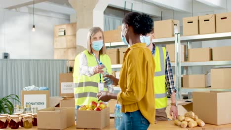 caucasian young male and senior female volunteers in facial mask giving food and water to homeless people in charity warehouse