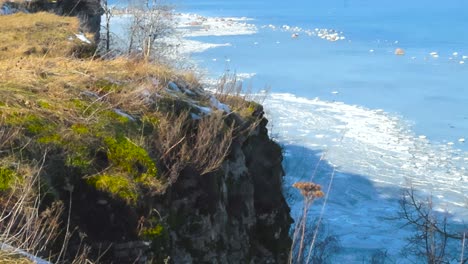 frozen blue baltic sea water during that has white ice and snow on it during a sunny day viewed from tabasalu high altitude cliff bank coastline during winter that has tall brown grass and moss on it.