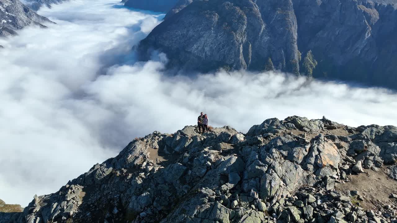 Drone orbits a couple standing at the edge of Bakkanosi cliffs, revealing the distant fjord below filled with fog