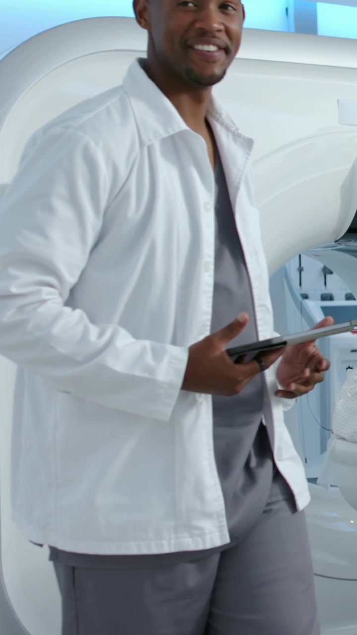 Video verticale: Medico in camice con tablet che istruisce un paziente in camice su un lettino da visita per una TAC.