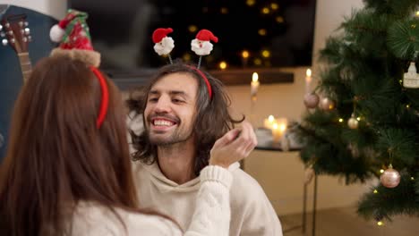 Over-the-shoulder,-a-brunette-girl-puts-on-her-brunette-boyfriend-in-a-white-sweatshirt-a-red-headband-with-Santa-Claus-emoticons-near-a-green-Christmas-tree-in-a-cozy-room-in-the-winter-evening