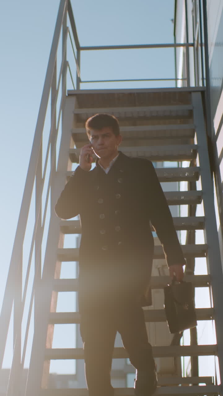 hombre con abrigo negro descendiendo la escalera en una llamada telefónica sosteniendo un maletín, asintiendo con la cabeza mientras habla bajo la luz del sol brillante, el fondo presenta vidrios reflectantes, coches estacionados y elementos urbanos