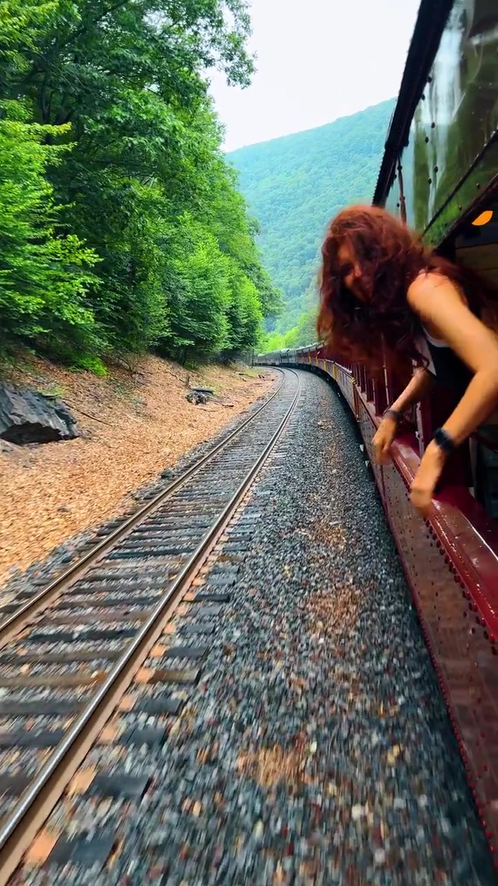 Passeggera che guarda fuori dal finestrino di un treno storico in movimento. Donna irriconoscibile che si sporge dal finestrino di un treno vintage rosso, osservando binari ferroviari curvi e montagne verdi lussureggianti.