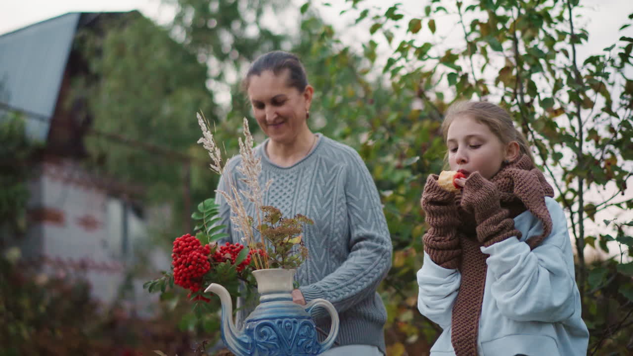Woman and girl arranging flowers outdoors, mother guiding daughter placing red berry bouquet into porcelain teapot vase on rustic porch near barn, scarves and sweaters signaling crisp autumn evening