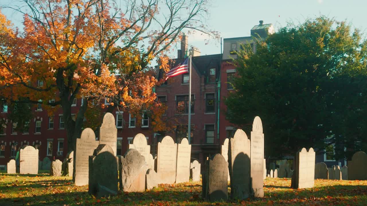 static shot of the Copps Hill Burying site during fall with tombstone in the foreground and the US flag in the background and leaves falling, Boston, Massachussetts, United States of America