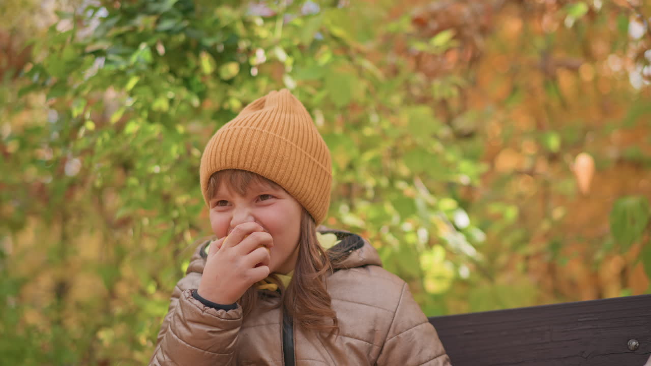 young girl in warm brown jacket and mustard beanie takes big bite of fresh apple while sitting on bench surrounded by vibrant autumn foliage, eyes lifted playfully as she enjoys sweet taste outdoors