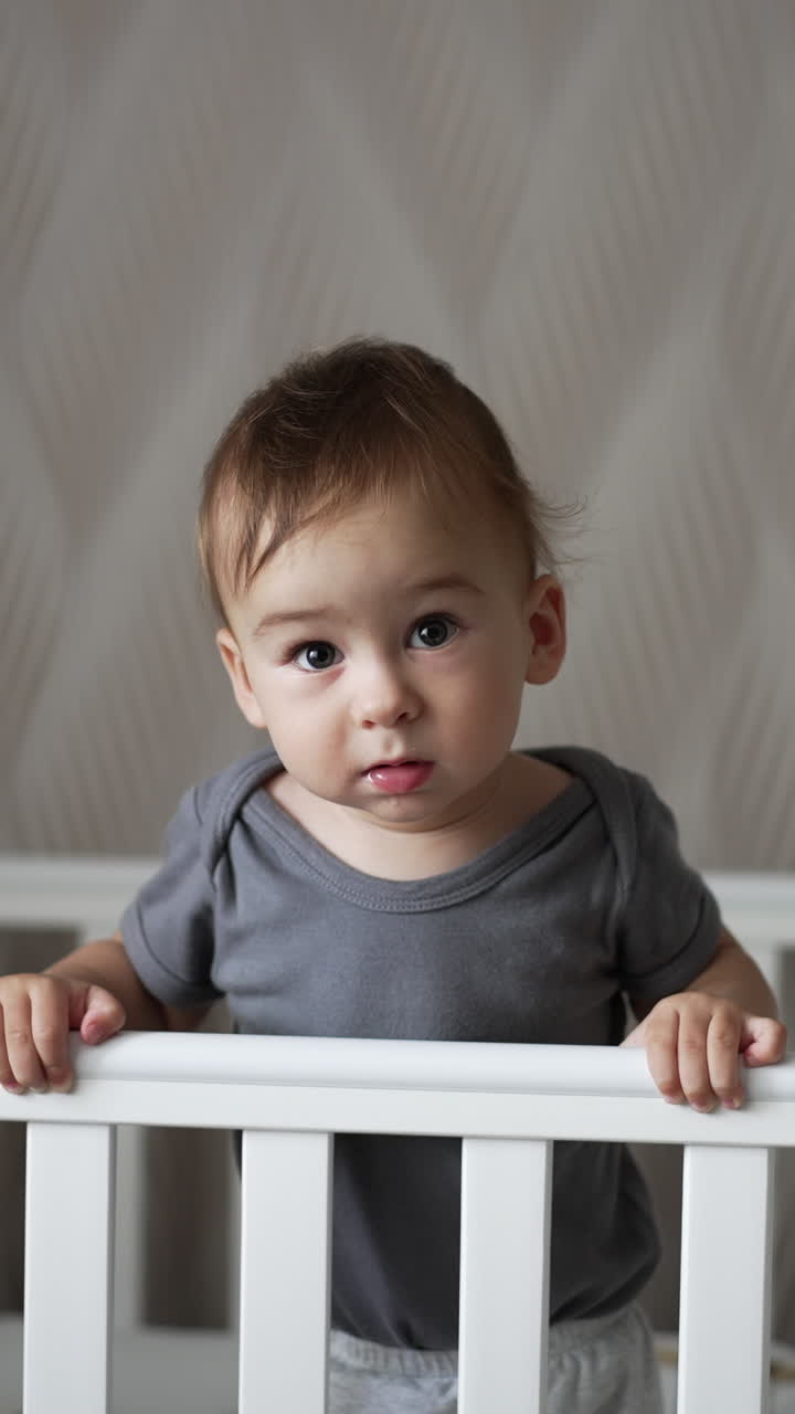 Calm Caucasian baby boy standing in a crib calmly. Child looks into camera and then smiles adorably. Light backdrop. Vertical video