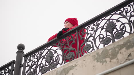 mujer con gorra roja y sudadera con capucha descansando las manos en intrincadas barandillas de hierro contra el cielo brillante de invierno, mostrando un elegante diseño de valla con una atmósfera serena y texturas sutiles de hormigón envejecido