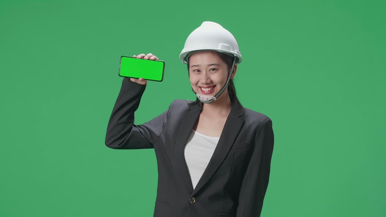 Smiling Asian Female Engineer With Safety Helmet Showing Green Screen Smartphone In The Green Screen Studio