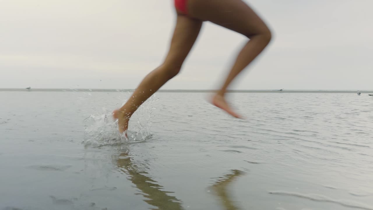 Young girl runs through shallow water at low tide, splashing under an overcast foggy sky, with seagulls wading along the beach shoreline