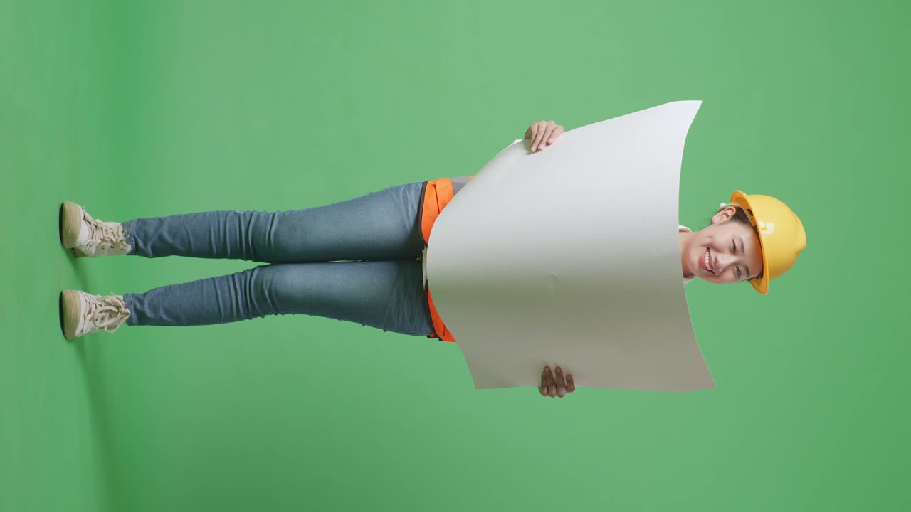 cuerpo lleno de ingeniera femenina asiática con casco de seguridad mirando el plano en sus manos y mirando a su alrededor mientras estaba de pie en el estudio de fondo de la pantalla verde