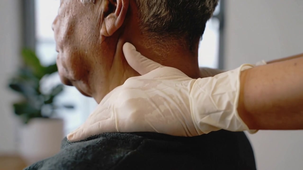 Un operatore sanitario che si avvicina e posa le mani guantate sul collo di un uomo anziano in clinica, valutando la tensione delle spalle.