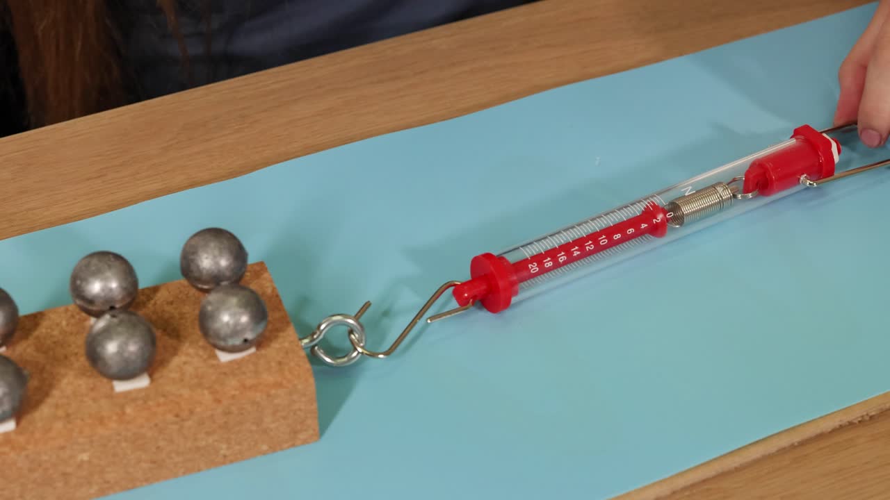 A hand pulls a weighted block using a spring scale on a smooth surface, illustrating friction and force dynamics