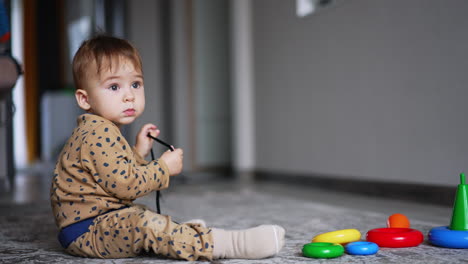 calm little baby sitting peacefully on the floor with a cord in hands. scattered parts of toy pyramid on the floor in front of child. blurred backdrop.