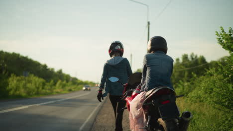 two women in helmets stand by the roadside with one seated on a motorcycle while the other walks away, in the background, cars approach from a distance on a quiet road