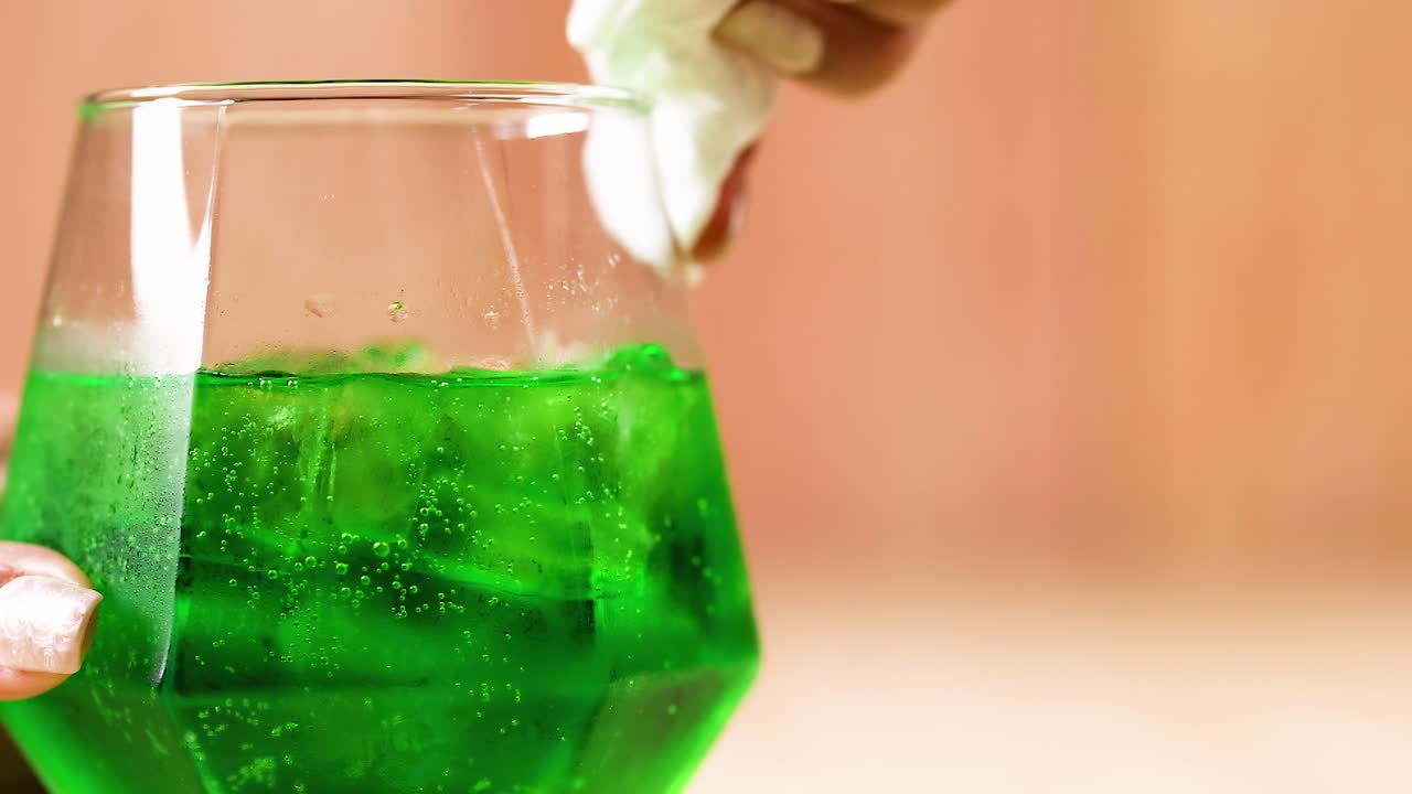A hand wipes the rim of a glass containing a vibrant green beverage.