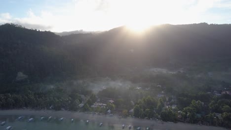 Aerial-of-small-fishing-village-by-the-beach-with-sun-rising-behind-hilltop-in-the-Philippines---camera-tracking-and-pedestal-down