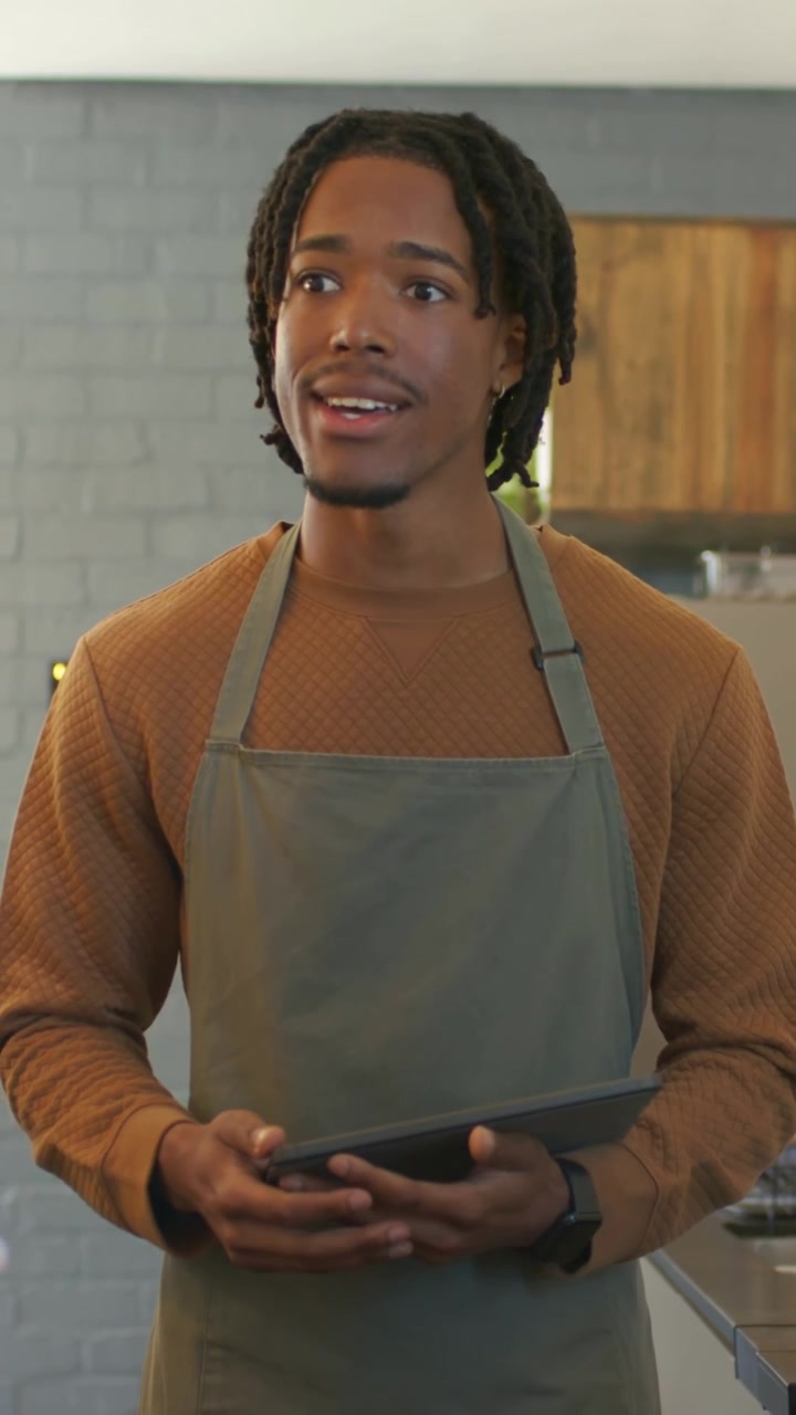Video verticale: Un cliente chiama fuori campo, mentre un barista con grembiule verde sistema il tablet al bancone del caffè.