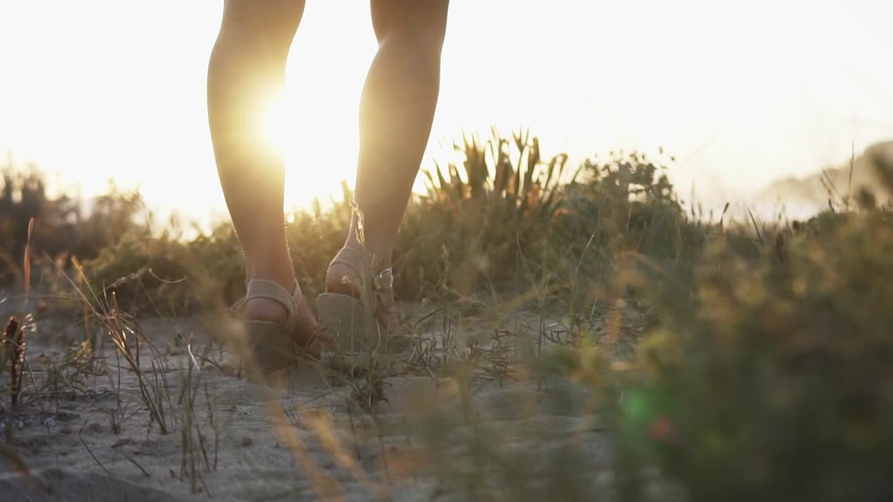 toma panorámica en cámara lenta de hermosas piernas de una mujer con tacones altos de pie en la playa rodeada de plantas y arena durante una hermosa puesta de sol