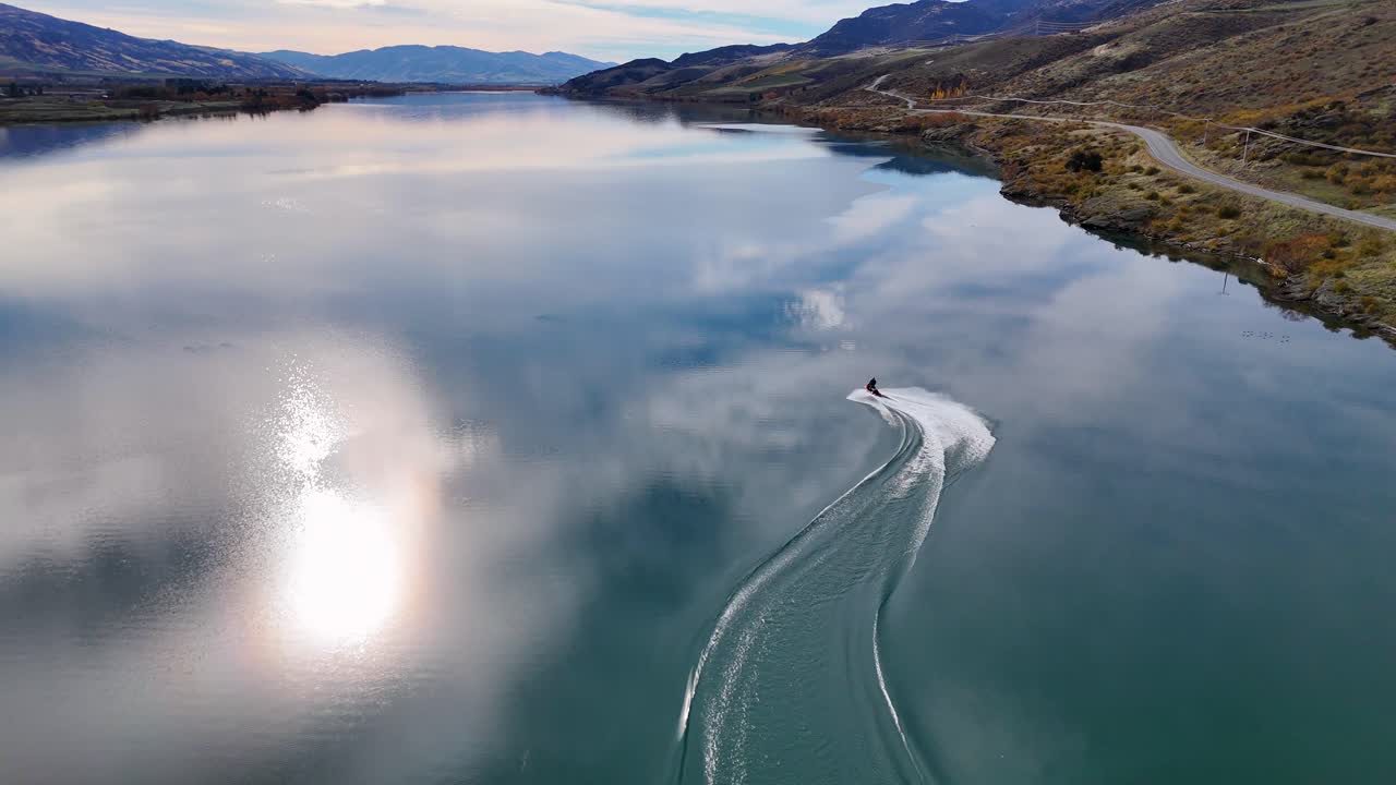 A jetski glides across Lake Dunstan, creating dynamic patterns on the water. Captured in Queenstown, New Zealand, under serene autumn lighting