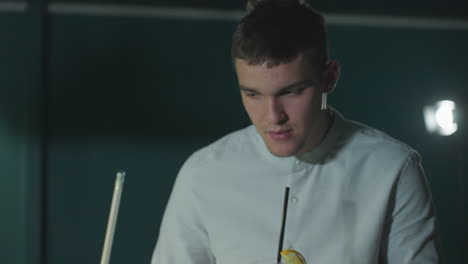 close-up of serious player in white shirt holding cue stick and sipping lemon drink with black straw while observing game. focused expression, ambient lighting, and modern billiards setting