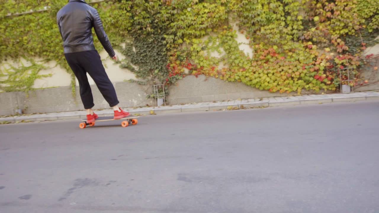 joven fresco en chaqueta de cuero patinando en su longboard en las calles