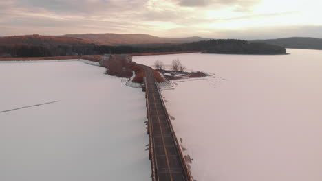 Luftflug-Direkt-über-Die-Architektonische-Brücke-Auf-Dem-Zugefrorenen-See-Im-Farbenfrohen-Abendlicht