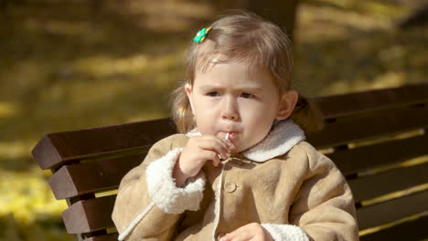 Entzückendes-Kleines-Mädchen,-Das-Lutscher-Isst,-Sitzt-Auf-Einer-Bank-Im-Herbstpark---Dolly-Von-Den-Beinen-Bis-Zum-Gesicht