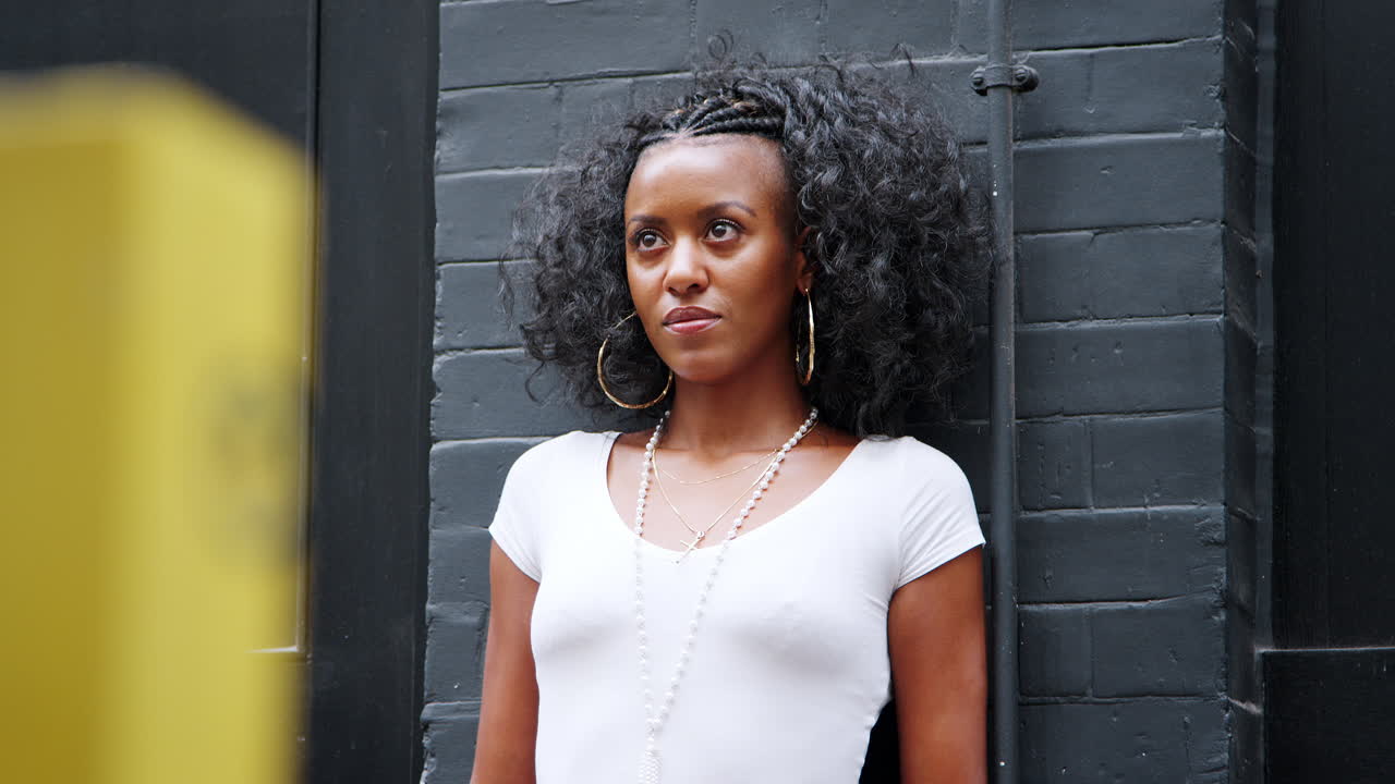 Mujer joven afroamericana elegante apoyada en una pared en la calle mirando al frente, de medio cuerpo