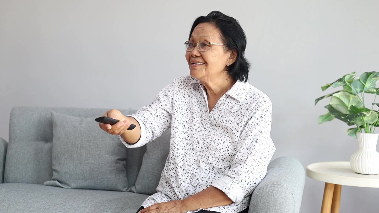 Felice donna anziana asiatica seduta sul divano in salotto, che punta il telecomando della TV per cambiare canale, guardando la sua serie di film online preferita.