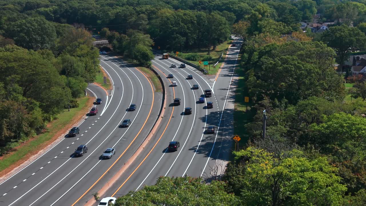 Aerial view of the Southern State Parkway on Long Island, NY during a sunny day with light automobile traffic. The camera dolly in and pan left over the trees standing next to the parkway