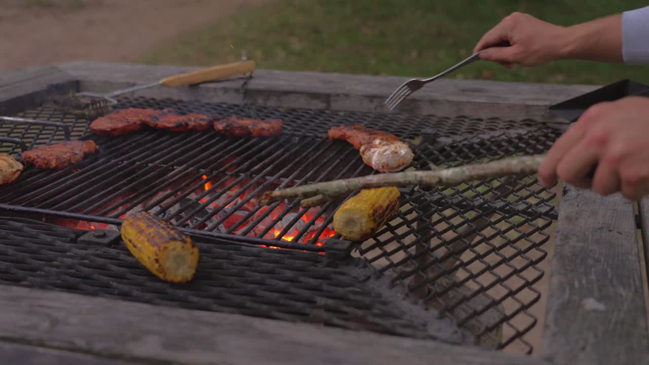 Grilling steak and corn on a rustic barbecue grill outdoors with a hand turning the food