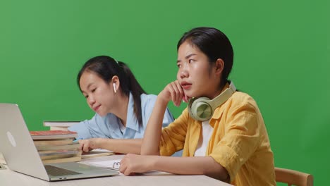 close up of asian woman student is bored while sitting with her friend and writing into the notebook on a table with a laptop in the green screen background classroom