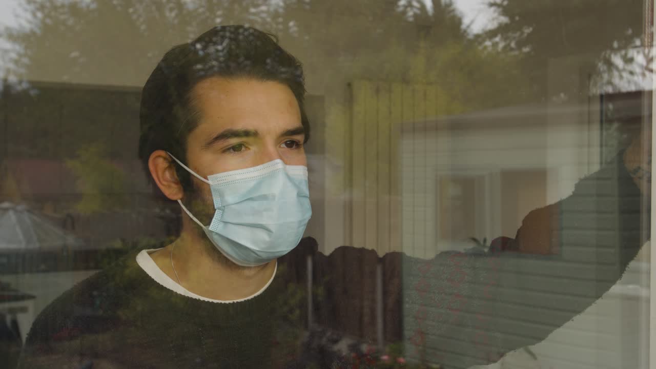 Male looking out and leaning on a window longingly wearing a facemask in quarantine