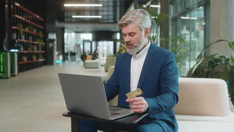 smiling businessman using credit card and laptop for online shopping payments in modern office lobby