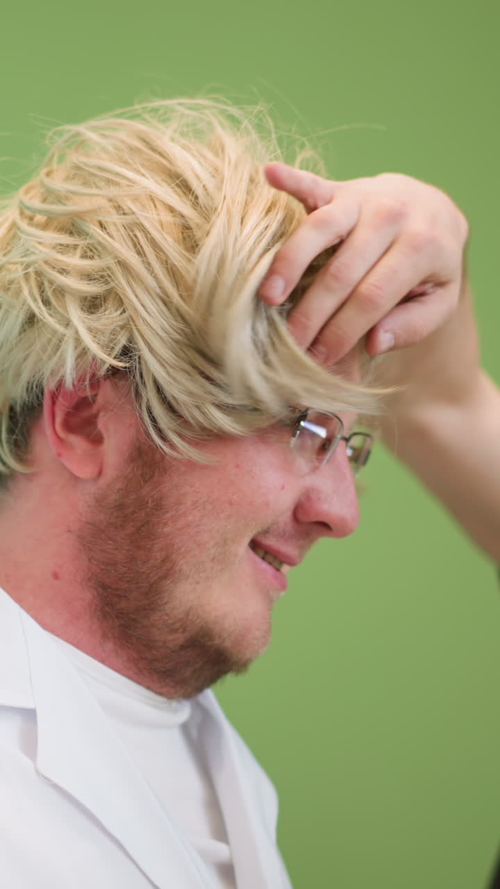 색 실험실 코트를 입은 남자가 금발의 위그와 검은색 장갑을 입고 있는 모습, 검은색 폴로 셔츠를 입은 다른 사람이 밝은 녹색 배경으로 위그를 장난스럽게 조정하는 모습