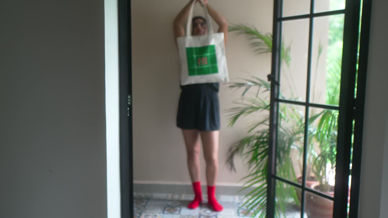 Giovane donna in piedi in un interno moderno che tiene una tote bag sopra la testa vicino a una finestra con luce naturale e piante da interno, creando una scena di stile di vita contemporaneo e rilassato