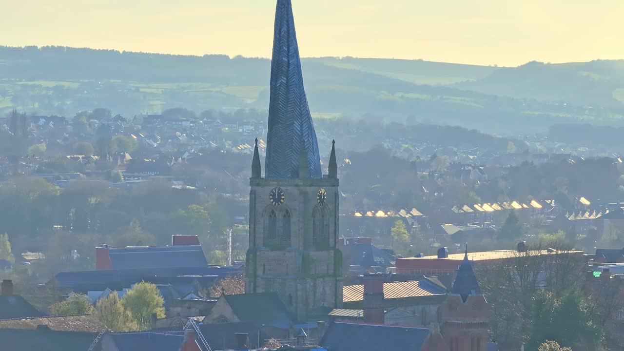 Aerial close-up of Chesterfield’s Cathedral Church, Derbyshire, England, focusing on its iconic twisted spire bathed in golden morning light, with rolling hills softly illuminated on the horizon.