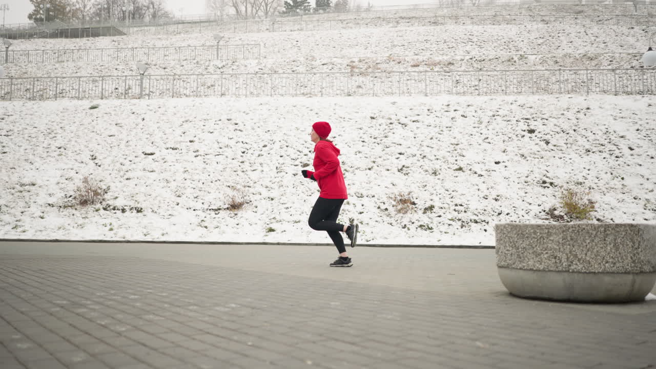 겨울 동안 눈 인 언덕 근처의 교차 도로에서 야외에서 조하는 성인의 측면 뷰, 빨간 후디를 입은 달리기 선수는 눈으로 여있는 언덕, 철 레일링을 특징으로하는 조용한 풍경에 활기찬 대조를 추가합니다.