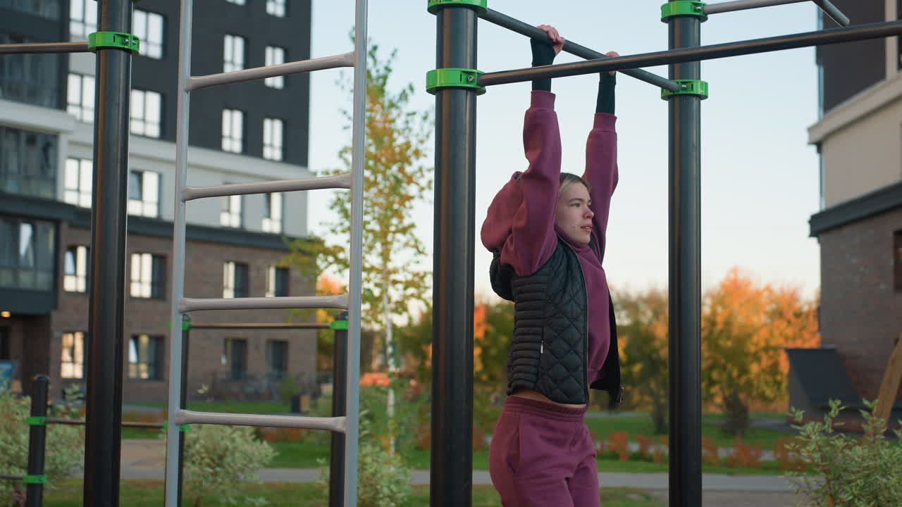 Side view of gym enthusiast gripping pull up bars while lifting legs in urban park workout, modern high rise backdrop, autumn foliage and children roller skating, focus on core strength and balance