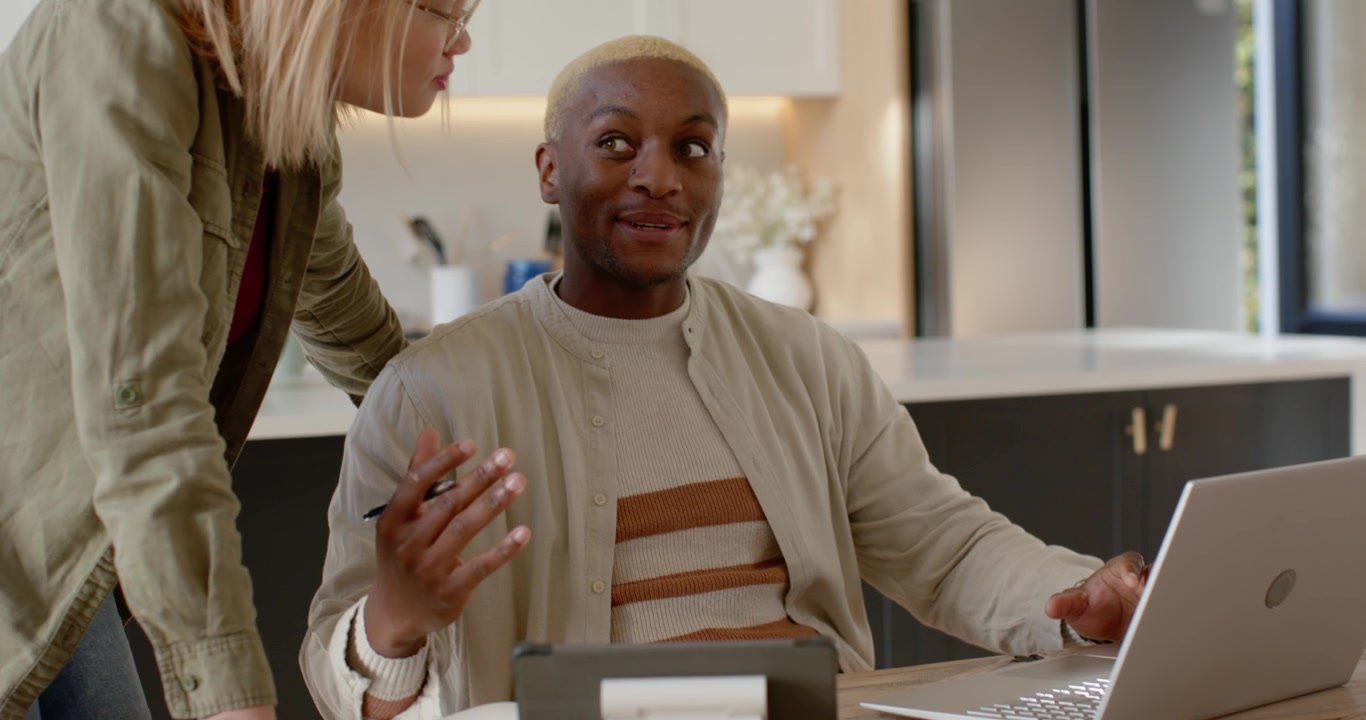 Diverse couple at kitchen table, woman leaning, man explaining, showing on laptop, phone reviewing
