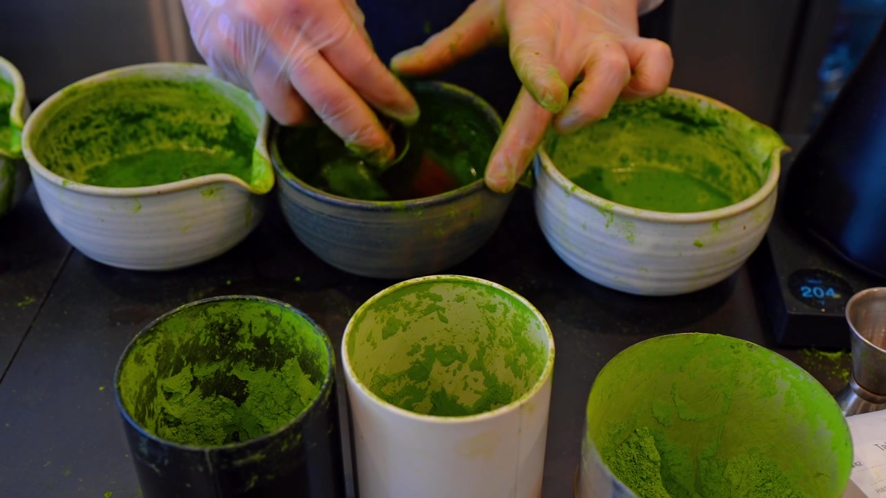 Close-up de pó de matcha em tigelas e recipientes em um bar de chá. Mãos enluvadas alcançam vasos de cerâmica revestidos com resíduo verde.