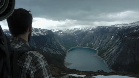 männlicher rucksacktourist auf dem berggipfel, der an einem bewölkten tag in norwegen einen atemberaubenden meerblick mit schnee auf dem gipfel genießt