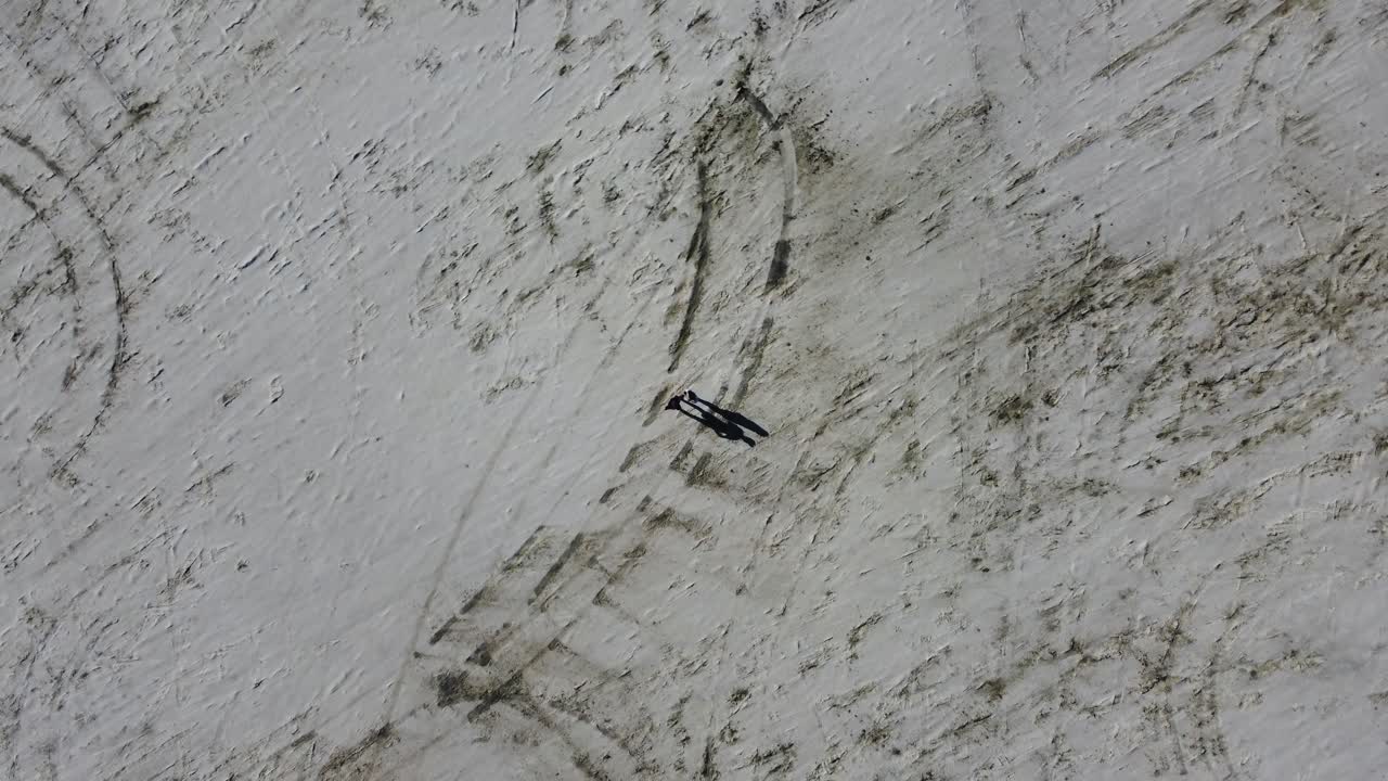 Slow zoom out aerial shot of two people standing on the Bonneville Salt Flats in the afternoon. This shot reveals the tire tracks of cars that have driven on the salt flats. Filmed with a drone in 4K.