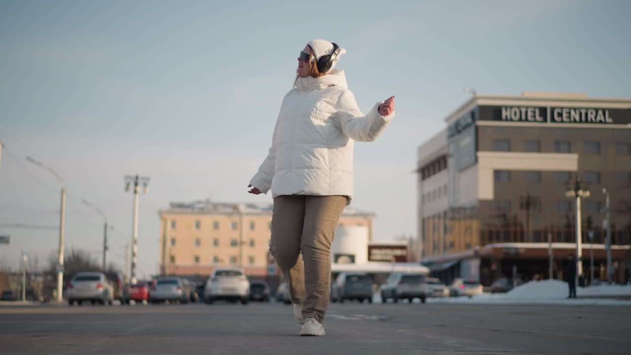 Performer dances confidently near car park wearing winter jacket and headphones, surrounded by snowy street and urban buildings, hotel sign visible in background, expressing joy through motion