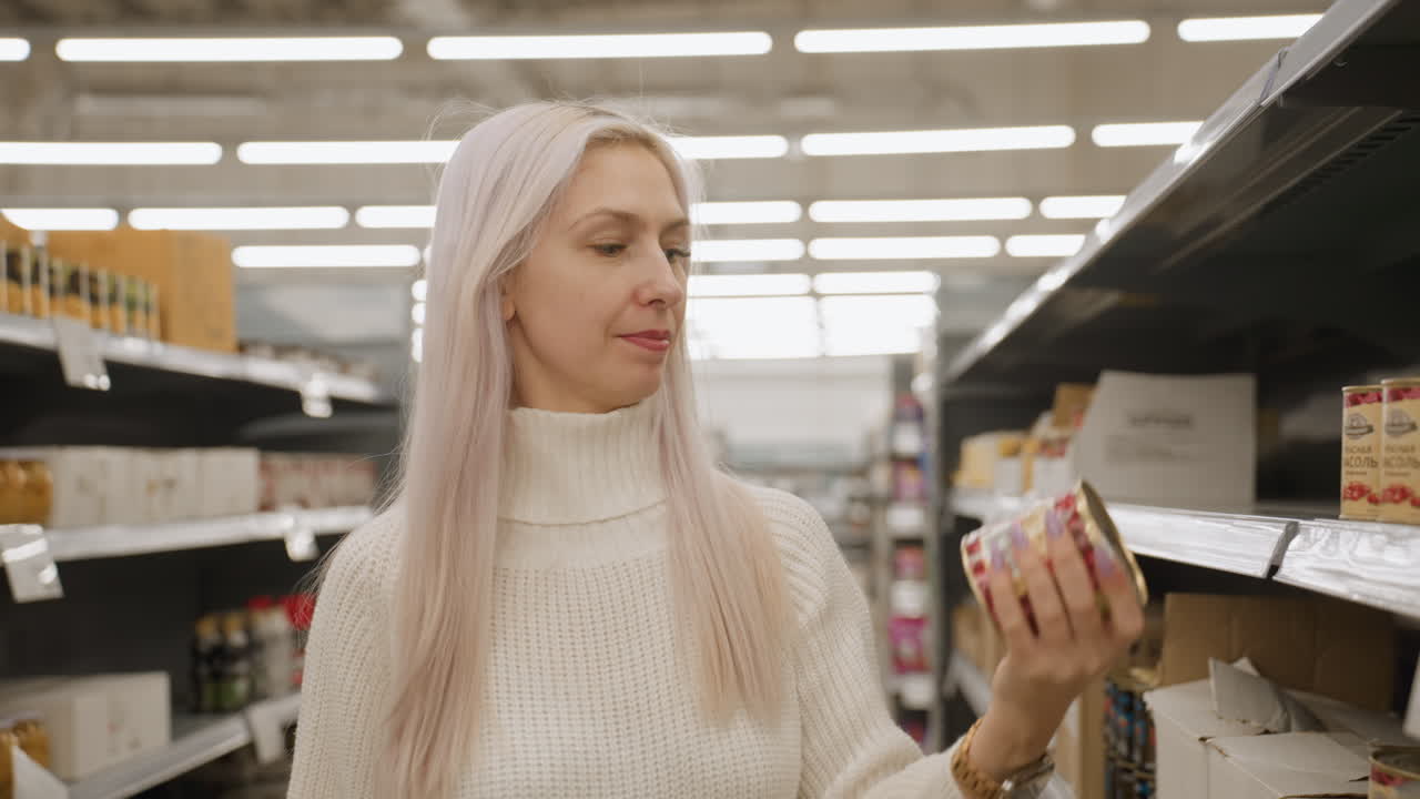Medium shot of house wife selecting canned food from supermarket shelf, tilts tin to examine expiry date printed on bottom under bright aisle lighting, weighing quality before placing into trolley